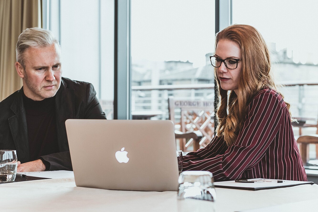 A man and a woman are sitting in front of a laptop together with the woman typing something on the laptop.